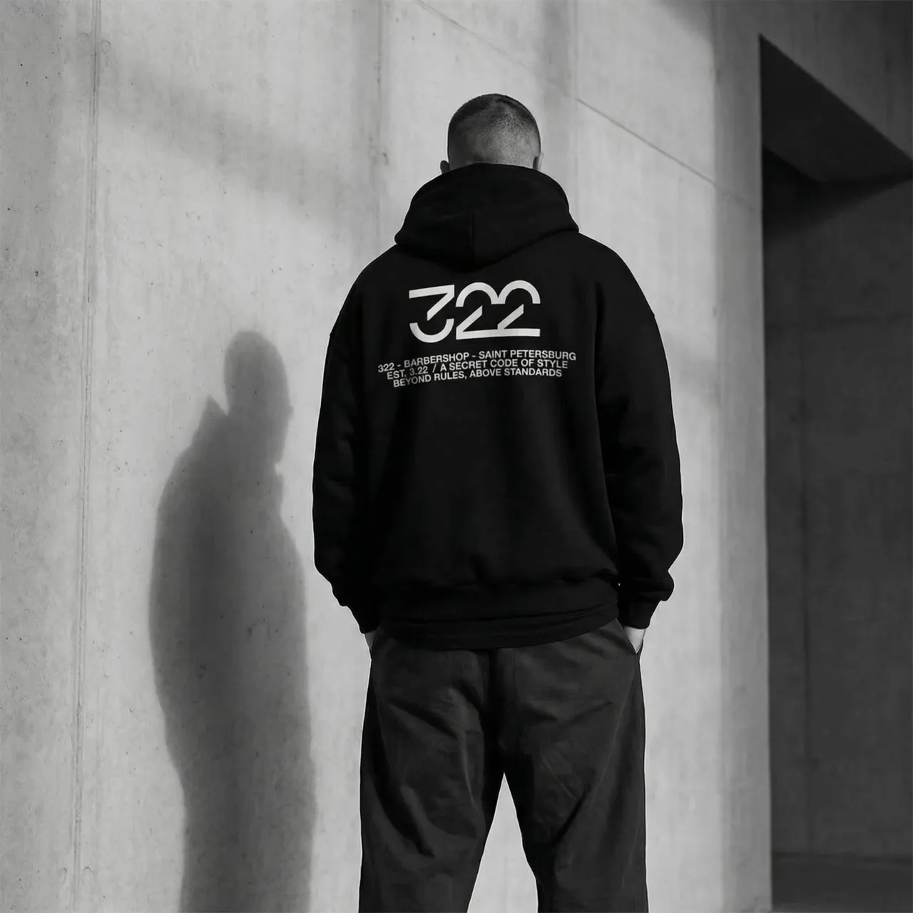 Subtle breathing motion of the person and hoodie, with a gentle, slow drift of soft light across the concrete wall. The shadow on the wall shifts slightly, creating a calm, premium atmosphere.